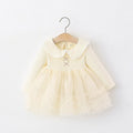 A pale yellow baby girl's dress with a ribbed long-sleeve bodice, a Peter Pan collar with white lace trim, and a layered tulle skirt, hanging on a wooden hanger against a white wall, Obroi