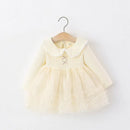 A pale yellow baby girl's dress with a ribbed long-sleeve bodice, a Peter Pan collar with white lace trim, and a layered tulle skirt, hanging on a wooden hanger against a white wall, Obroi