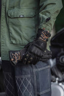 Black motorcycle leather glove held by a person wearing green pants, with promotional text on the right, Obroi