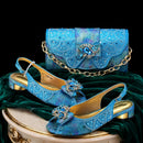 A matching blue embellished shoe and handbag set displayed on a dark green velvet drape over a gold-trimmed pedestal against a black background. The set includes two pointed-toe slingback flats with jeweled bows and heels, and a structured clutch with a gold chain strap and jeweled floral motif, Obroi