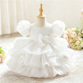 White toddler girl's party dress displayed on a dress form mannequin, featuring puff sleeves, a large central bow with a rose applique, and a tiered ruffled skirt with matching rose details, Obroi