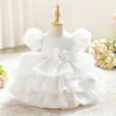 White toddler girl's party dress displayed on a dress form mannequin, featuring puff sleeves, a large central bow with a rose applique, and a tiered ruffled skirt with matching rose details, Obroi