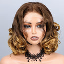 A shoulder-length wavy wig with dark brown roots and golden blonde highlights, styled on a mannequin head with a black lace choker against a light gray background. The brand name 'Obroi' is written in black script in the top left corner, Obroi