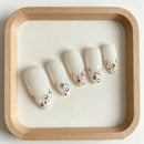 Five white press-on nails with colorful rhinestone embellishments, arranged on a white insert inside a light wood tray Obroi