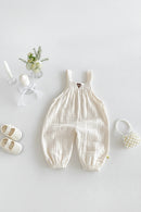A baby's cream-colored crinkle gauze overall romper with elastic cuffs, displayed in a styled flat-lay with white baby shoes, a pearl headband, a small flower arrangement, and candles on a light gray background, Obroi