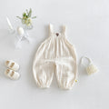 A baby's cream-colored crinkle gauze overall romper with elastic cuffs, displayed in a styled flat-lay with white baby shoes, a pearl headband, a small flower arrangement, and candles on a light gray background, Obroi