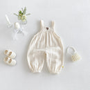 A baby's cream-colored crinkle gauze overall romper with elastic cuffs, displayed in a styled flat-lay with white baby shoes, a pearl headband, a small flower arrangement, and candles on a light gray background, Obroi