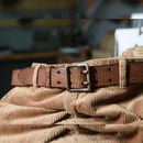 A close-up of a brown leather belt with a square metal buckle resting on a ribbed tan corduroy fabric, Obroi