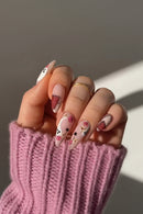 Hand with intricately designed nails featuring flowers and polka dots on a pink textured surface, Obroi