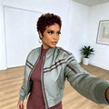 A woman taking a mirror selfie wearing a sage green bomber jacket with brown stripes over a ribbed mauve-brown dress, in a bright room with light wood floors and white walls, Obroi