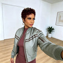 A woman taking a mirror selfie wearing a sage green bomber jacket with brown stripes over a ribbed mauve-brown dress, in a bright room with light wood floors and white walls, Obroi