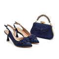 Navy blue high-heeled shoes with floral details and a matching handbag on a white background, Obroi