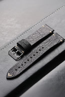 Black leather watch strap with silver buckle on a gray surface, Obroi