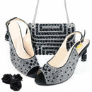 A coordinated women's accessory set on a white background: a pair of black peep-toe slingback heels covered in silver rhinestones, a matching black handbag with silver rhinestone and bead detailing, and three black fabric roses in the lower left, Obroi