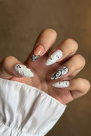 A close-up of a hand showcasing Halloween-themed nail art. The nails are predominantly white with black details, featuring ghost and mummy designs, with one accent nail painted in a soft peach color and another decorated with a black-and-white patterned lace-like design, Obroi