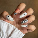 A close-up of a hand showcasing Halloween-themed nail art. The nails are predominantly white with black details, featuring ghost and mummy designs, with one accent nail painted in a soft peach color and another decorated with a black-and-white patterned lace-like design, Obroi