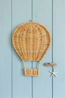 A natural woven wicker hot air balloon wall hanging with a small metal hook at the top, displayed against a light blue painted wooden background with a small driftwood piece and starfish decoration nearby, Obroi
