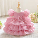 A pink toddler girl's party dress displayed on a small mannequin dress form. The dress features a tiered ruffle skirt, puffy sleeves, and a large bow with a rose at the center, accented with fabric roses on the sides, Obroi
