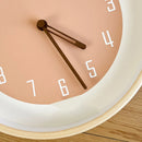 Close-up of a minimalist wall clock with a warm peach-pink face, white numerals, dark brown hands, and a light wood rim, resting on a light wood surface, Obroi