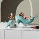 Two whale-shaped decorative sculptures on a white cabinet, each with a small human figure posed on top. The larger whale is mint green with a white underside and a figure reclining on it, while the smaller whale is pale blue and white with a figure sitting on top, Obroi