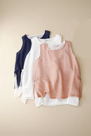 Three sleeveless linen-style tank tops in navy blue, white, and dusty pink, layered flat on a light beige background with a small magazine corner visible in the top right, Obroi