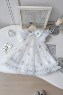 A white toddler girl's dress with a ruffled Peter Pan collar, decorated with embroidered silver and gold stars, displayed on a hanger on a white table with decorative props, Obroi