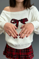 Person wearing a white sweater with a red plaid bow design, holding hands in a heart shape, Obroi