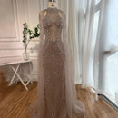 A floor-length nude-beige evening gown with intricate silver beadwork and a matching sheer tulle overskirt, displayed on a mannequin in a bright room with white curtains and wood flooring, Obroi