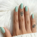 Hand with light green nails and subtle tattoos on a soft white background, Obroi
