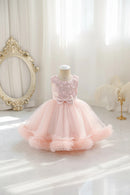 A toddler girl's formal party dress displayed on a mannequin in a bright, elegant room. The dress features a light pink sequined bodice with a large bow at the waist and a voluminous, layered tulle skirt in pale pink, Obroi