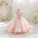 A toddler girl's formal party dress displayed on a mannequin in a bright, elegant room. The dress features a light pink sequined bodice with a large bow at the waist and a voluminous, layered tulle skirt in pale pink, Obroi