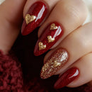 Red glittery nails with gold heart and white feather designs on a dark background, Obroi