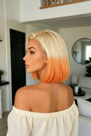 A woman shown from the back and side profile wearing a short bob haircut with a blonde top fading into a bright orange ombre at the ends, photographed indoors in a modern living room, Obroi