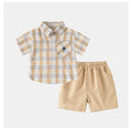A two-piece baby boy's outfit set featuring a short-sleeve plaid button-up shirt in white, yellow, and gray, paired with beige elastic-waist shorts, displayed on a light gray background, Obroi