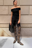 Person wearing a black outfit with knee-high brown boots standing against a textured wall, Obroi