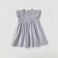 A light gray baby girl's dress with ruffled cap sleeves and vertical smocked pleats on the bodice, laid flat against a light gray background, Obroi