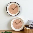 Two round wall clocks with dusty pink faces and white rims, displayed on a light wall above a wooden surface with a wicker basket and green leaves, Obroi