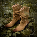 Pair of brown suede boots on a natural background with green foliage, Obroi