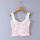A light pink floral sleeveless top with white lace shoulder straps and a white front zipper, hanging on a wooden hanger against a light gray background, Obroi