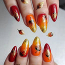 Close-up of manicured fingernails with a fall-themed design. The nails feature almond-shaped tips with glossy red and orange-to-yellow gradient tips, decorated with metallic gold foil leaf designs and a few black leaf accents, Obroi
