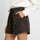 A person wearing high-waisted leopard-print shorts with a black drawstring waistband, paired with a white long-sleeve top, shown against a light gray studio background, Obroi