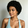 A woman with a short, curly black wig wearing a light blue tank top and gold hoop earrings, shown from the chest up against a plain light gray background, Obroi
