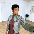 A woman taking a mirror selfie wearing a light sage green satin bomber jacket with brown stripes over a dusty rose ribbed dress, in a bright room with light wood floors, Obroi