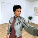 A woman taking a mirror selfie wearing a light sage green satin bomber jacket with brown stripes over a dusty rose ribbed dress, in a bright room with light wood floors, Obroi