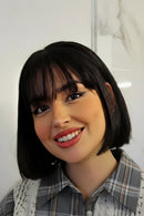 A smiling woman with a short black bob haircut and straight bangs, wearing a plaid shirt, set against a light marble wall background, Obroi