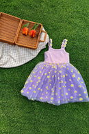A toddler girl's sleeveless dress laid flat on green grass, featuring a light pink ribbed bodice and a voluminous lavender tulle skirt printed with small white daisies with yellow centers. A wicker picnic basket, orange tulips, and a beige-and-white gingham blanket are styled around it, Obroi