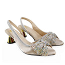 A pair of champagne satin slingback pumps with crystal-embellished bows and metallic gold block heels, shown on a white background with a 'Obroi' logo in the top left corner, Obroi