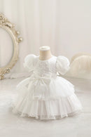 A white baby girl's formal dress displayed on a cream mannequin dress form. The dress features puffy short sleeves, a lace-trimmed bodice, a large central bow, and a tiered tulle skirt, Obroi