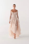 A model wearing a pale blush pink lace and tulle midi dress with long sheer sleeves, a sweetheart neckline, and a layered high-low skirt, photographed against a seamless light gray studio background.