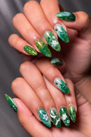 Hand with green and white floral nail art on a neutral background, Obroi